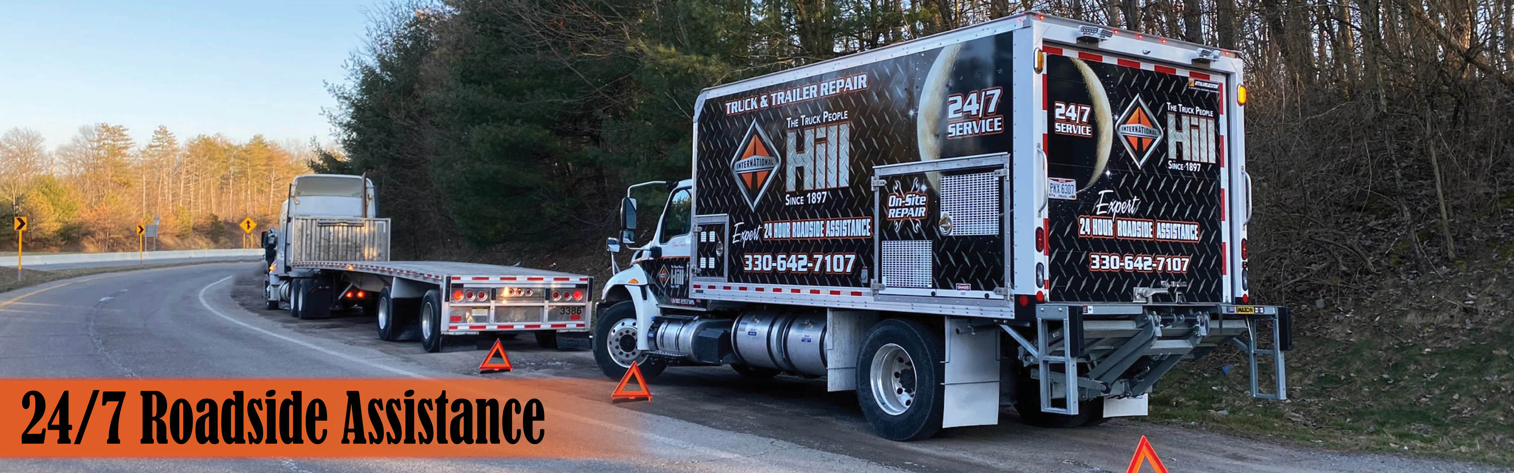 Truck with an open bed pulled over on the side of a road next to a 24-Hour Roadside Assistance truck.