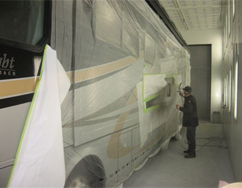 Truck body shop technicians wearing black working on a black & yellow-striped RV with film over it.