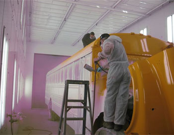 Two body repair technicians painting a bus white inside a garage with white walls and ceiling.